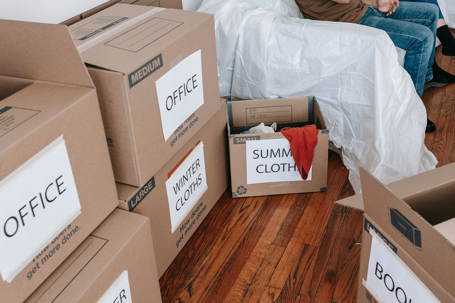 A couple seated on a couch surrounded by labeled moving boxes in a bright room, preparing to relocate.